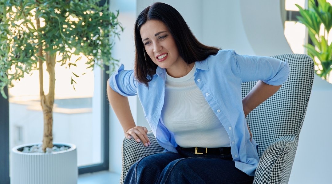 Woman experiencing lower back pain while sitting in a chair and holding her lower back.